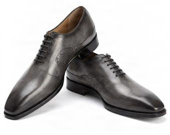 May include: A pair of grey leather oxford shoes featuring a burnished finish, black laces, and intricate embossed patterns on the side panels. The shoes have a pointed toe, a low stacked heel, and a smooth, polished surface with dark shading.