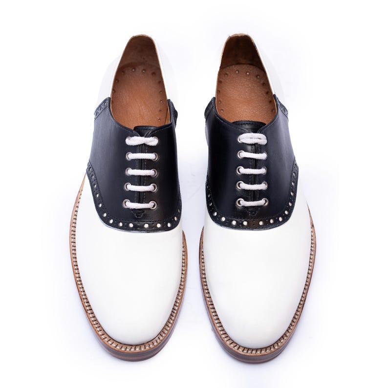 May include: A pair of two-tone saddle shoes. The shoes have a white vamp and a black saddle and heel. White laces thread through the black leather. The soles are brown with a stitched border. The shoes are displayed on a white background.