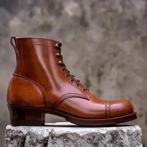 May include: A brown leather boot with a cap toe and a stacked heel. The boot has a lace-up closure with brass eyelets and a pull tab at the top. The leather has a polished finish. The boot is approximately 6 inches tall.