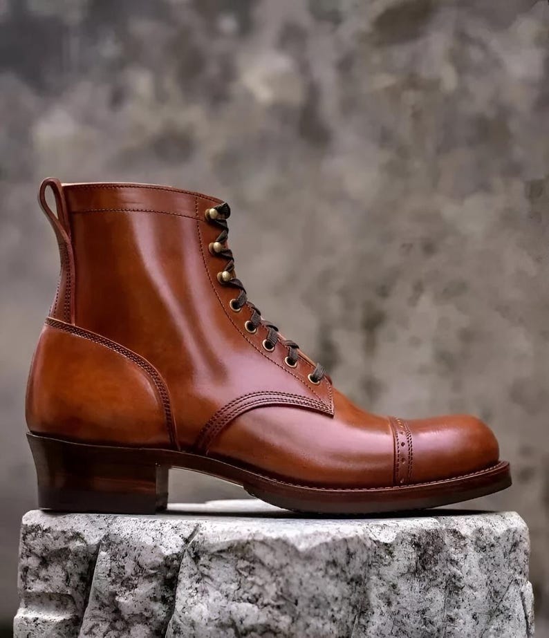 May include: A brown leather boot with a cap toe and a stacked heel. The boot has a lace-up closure with brass eyelets and a pull tab at the top. The leather has a polished finish. The boot is approximately 6 inches tall.