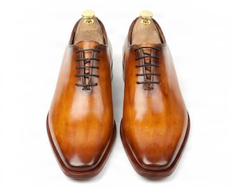 May include: A pair of tan leather oxford shoes with a polished finish, featuring dark brown laces and a closed lacing system. Each shoe includes a wooden shoe tree with a gold-colored metal knob inserted into the opening for shape retention.