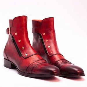 Könnte beinhalten: Ein Paar Stiefeletten in tiefroter Ausführung mit strukturierter Oberfläche. Jeder Stiefel verfügt über einen seitlichen Reißverschluss, einen dekorativen Riemen mit Metallschnalle und eine Reihe kleiner Metallnieten entlang des Reißverschlusses, auf einem niedrigen Blockabsatz.
