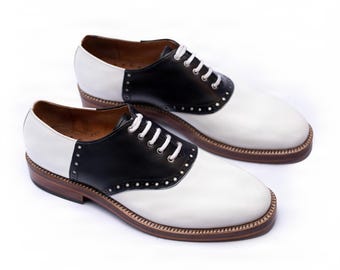 May include: A pair of saddle shoes featuring a two-tone design with white leather on the toe and heel, black leather on the midsection, white laces, and a brown stacked heel with a welted sole. The interior lining is a light brown leather.