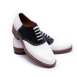 May include: A pair of saddle shoes with white leather uppers and black leather saddle panels. The shoes have white laces, brown soles, and decorative stitching around the edges. A classic, preppy style.
