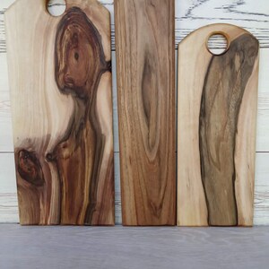 Op de afbeelding: Drie rechthoekige houten serveerplanken met afgeronde bovenkanten en ronde uitsparingen om op te hangen. Elke plank heeft een unieke natuurlijke houtnerf met vari&euml;rende tinten van licht cr&egrave;me, beige en donkerbruin met organische knoesten.