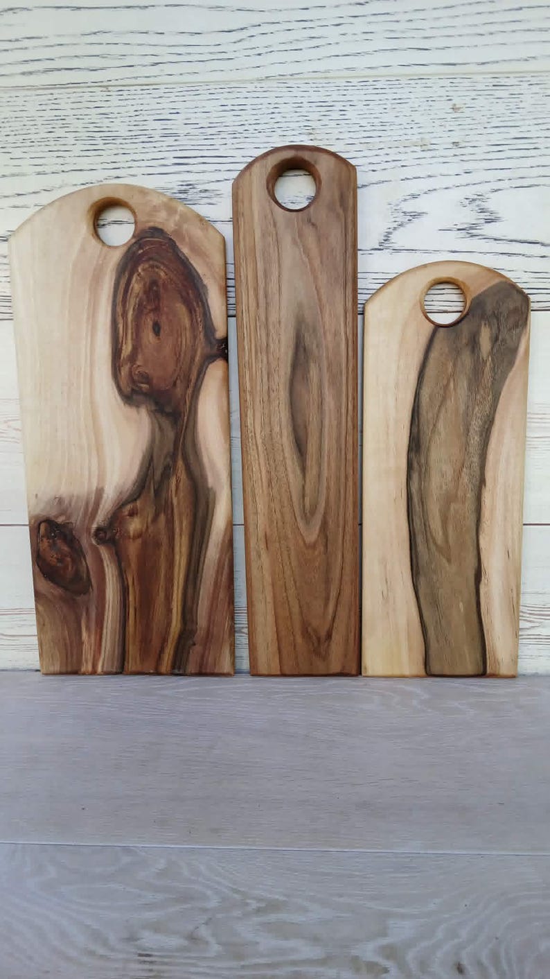 Op de afbeelding: Drie rechthoekige houten serveerplanken met afgeronde bovenkanten en ronde uitsparingen om op te hangen. Elke plank heeft een unieke natuurlijke houtnerf met vari&euml;rende tinten van licht cr&egrave;me, beige en donkerbruin met organische knoesten.