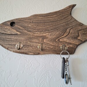 May include: A wooden key holder shaped like a fish, with a dark brown finish and visible wood grain. It features three metal hooks for keys and a key ring with keys attached. The fish design includes a circular eye and a curved mouth.