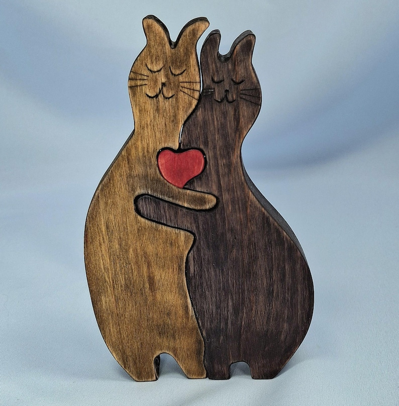 May include: A wooden cat ornament in shades of brown, holding a red heart. The cats are carved with closed eyes and whiskers, creating a loving scene. A unique decorative item for cat lovers.