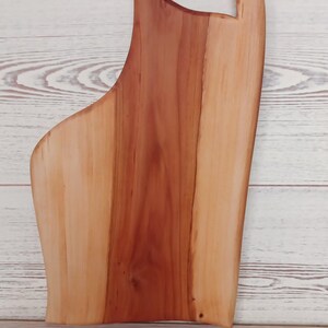 Puede incluir: Una tabla de servir de madera con un borde natural y un acabado liso y pulido. La tabla muestra una clara variaci&oacute;n de color entre el centro de color marr&oacute;n rojizo intenso y los bordes de color beige claro, con un asa integrada en la parte superior.