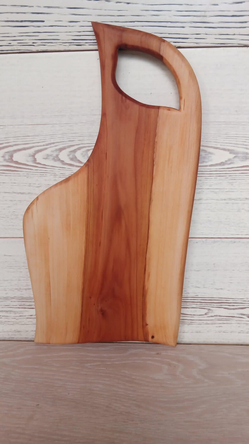 Puede incluir: Una tabla de servir de madera con un borde natural y un acabado liso y pulido. La tabla muestra una clara variaci&oacute;n de color entre el centro de color marr&oacute;n rojizo intenso y los bordes de color beige claro, con un asa integrada en la parte superior.