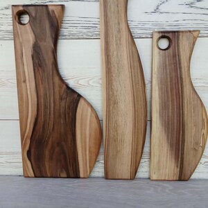 Op de afbeelding: Drie houten serveerplanken met natuurlijke houtnerven en vari&euml;rende breedtes. Elke plank heeft een glad, gebogen zijprofiel en een rond gat aan de bovenkant om op te hangen. Het hout vertoont een mix van lichte en donkerbruine tinten.