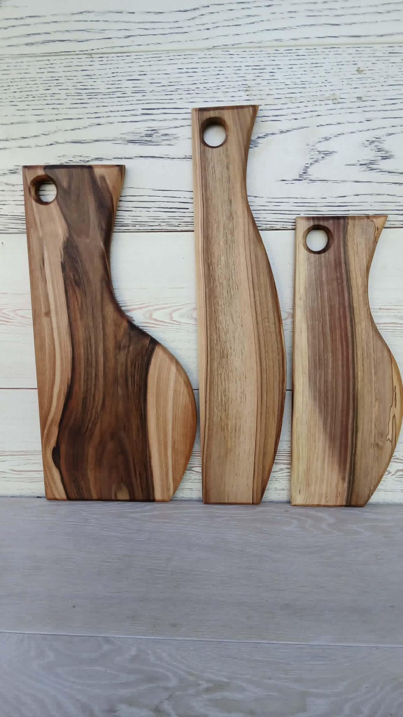 Op de afbeelding: Drie houten serveerplanken met natuurlijke houtnerven en vari&euml;rende breedtes. Elke plank heeft een glad, gebogen zijprofiel en een rond gat aan de bovenkant om op te hangen. Het hout vertoont een mix van lichte en donkerbruine tinten.