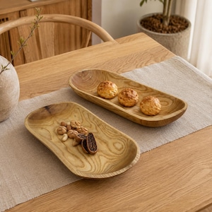 Op de afbeelding: Twee ovale houten schalen met een zichtbaar natuurlijk nerfpatroon. E&eacute;n schaal bevat drie ronde, goudbruine gebakjes, terwijl de andere schaal een kleine selectie gemengde noten bevat, waaronder amandelen en stukjes gedroogd fruit.