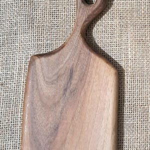 May include: A rectangular wooden cutting board with a smooth, light brown finish and visible natural wood grain patterns. The board features a rounded handle at the top with a circular hole for hanging, and a flat, straight base for stability.