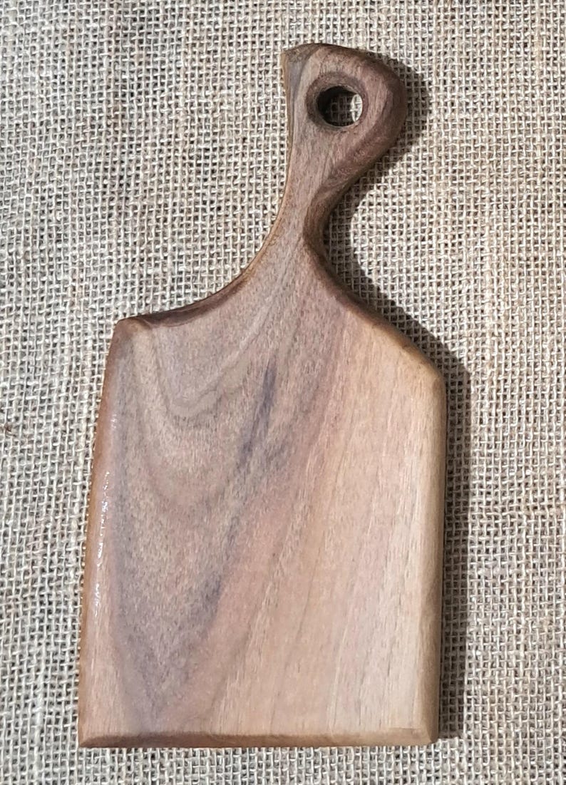 May include: A rectangular wooden cutting board with a smooth, light brown finish and visible natural wood grain patterns. The board features a rounded handle at the top with a circular hole for hanging, and a flat, straight base for stability.
