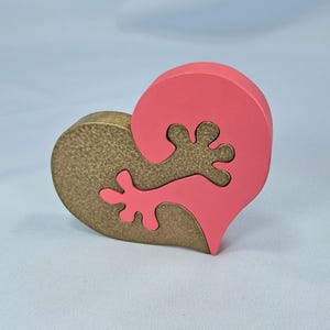以下が含まれることがあります： 木製のハート型のパズル。ベースは金色で、上部はコーラル色です。上部はベースに収まる有機的な形状をしています。パズルは連動するように設計されています。