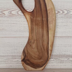 May include: A curved wooden serving board featuring a natural grain pattern with dark brown and light tan tones. The board has an elongated, organic shape with a circular cutout near the top, serving as a handle for easy gripping and carrying.