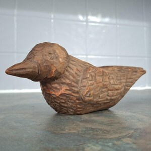 Pu&ograve; includere: Scultura di uccello in legno intagliato a mano. L'uccello &egrave; marrone con piume intagliate dettagliate e un becco prominente. La scultura &egrave; appoggiata su una superficie grigia, con uno sfondo piastrellato bianco.