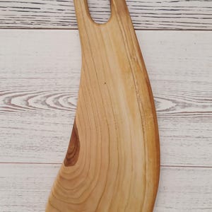 May include: A light brown wooden serving board with a curved, elongated shape and a smooth, polished finish. The board features a prominent natural wood grain pattern and a single oval-shaped handle cutout at the top, showcasing the organic texture.