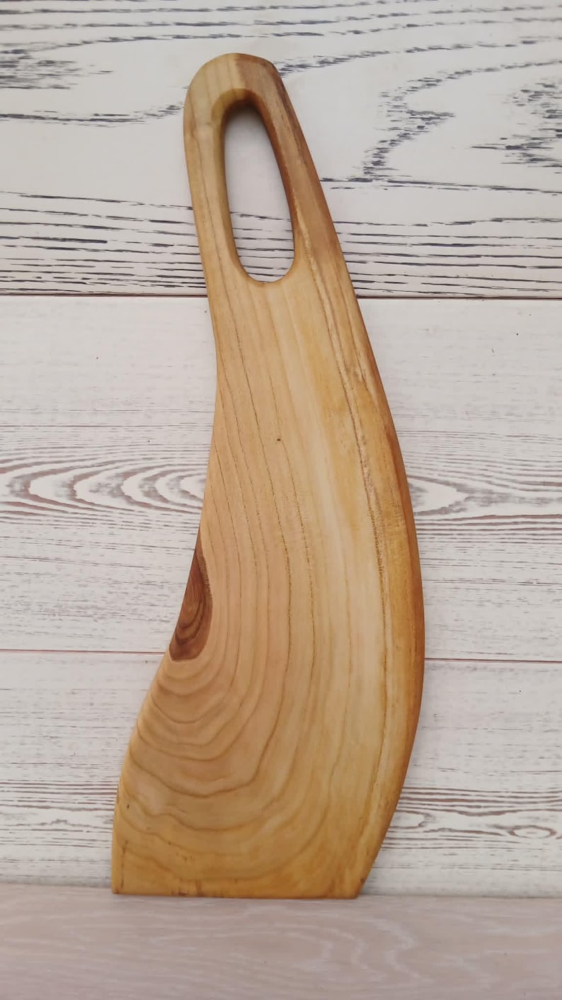 May include: A light brown wooden serving board with a curved, elongated shape and a smooth, polished finish. The board features a prominent natural wood grain pattern and a single oval-shaped handle cutout at the top, showcasing the organic texture.