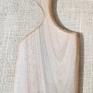 May include: A light brown wooden serving board with a smooth, flat rectangular body and a curved handle featuring a circular hole at the top. The wood displays a natural grain pattern with subtle variations in tone across the surface.