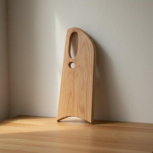 May include: A light-colored wooden serving board with a smooth, natural grain finish. The board features an asymmetrical, elongated shape with a curved bottom edge and two distinct circular and oval cutouts near the top for handling.
