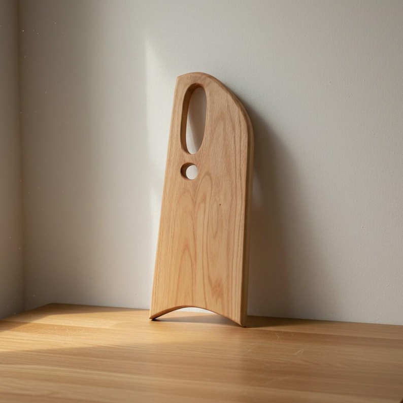 May include: A light-colored wooden serving board with a smooth, natural grain finish. The board features an asymmetrical, elongated shape with a curved bottom edge and two distinct circular and oval cutouts near the top for handling.