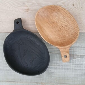 K&ouml;nnte beinhalten: Zwei Serviertabletts aus Holz. Eines ist schwarz gebeizt, das andere Naturholz. Beide haben eine ovale Form mit einem Griff mit einem Loch zum Aufh&auml;ngen. Die Tabletts werden auf einer hellen Holzoberfl&auml;che pr&auml;sentiert.