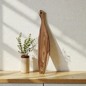 May include: A tall, narrow wooden serving board with a natural grain pattern and a small hole at the top of the handle, positioned vertically next to a cylindrical cream-coloured ceramic vase containing a small green leafy plant.