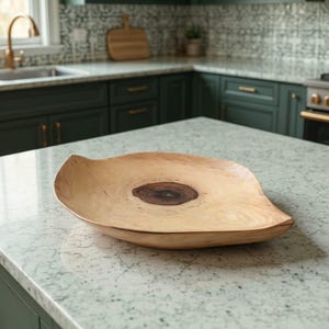 Puede incluir: Un gran cuenco de madera hecho a mano con una forma natural y orgánica. El cuenco presenta una madera de color claro con un nudo oscuro y circular en el centro. Los bordes se curvan suavemente hacia arriba, creando un plato poco profundo.