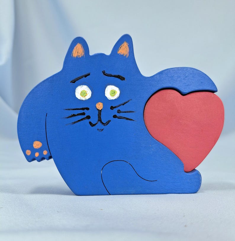 以下が含まれることがあります： 赤いハートを抱えた青い木製の猫の置物。猫はオレンジ色の耳と足パッドがあり、緑の目と黒いひげが描かれた顔をしています。ハートは猫が抱きしめる別の部分です。