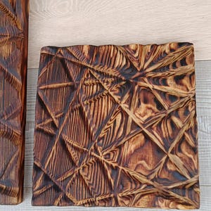 May include: A square, wooden decorative piece with a geometric, carved design. The wood has a dark brown, burnt finish, with a textured surface. The piece is placed on a light wood surface.