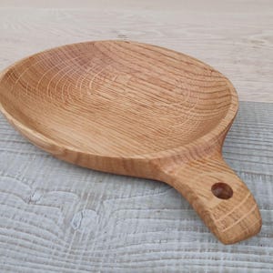 K&ouml;nnte beinhalten: Ein hellbraunes, ovales Serviertablett aus Holz mit Griff. Das Tablett hat eine flache Schale und einen Griff mit einem Loch zum Aufh&auml;ngen. Die Holzmaserung ist &uuml;ber das gesamte Tablett sichtbar, das etwa 25 cm lang ist.