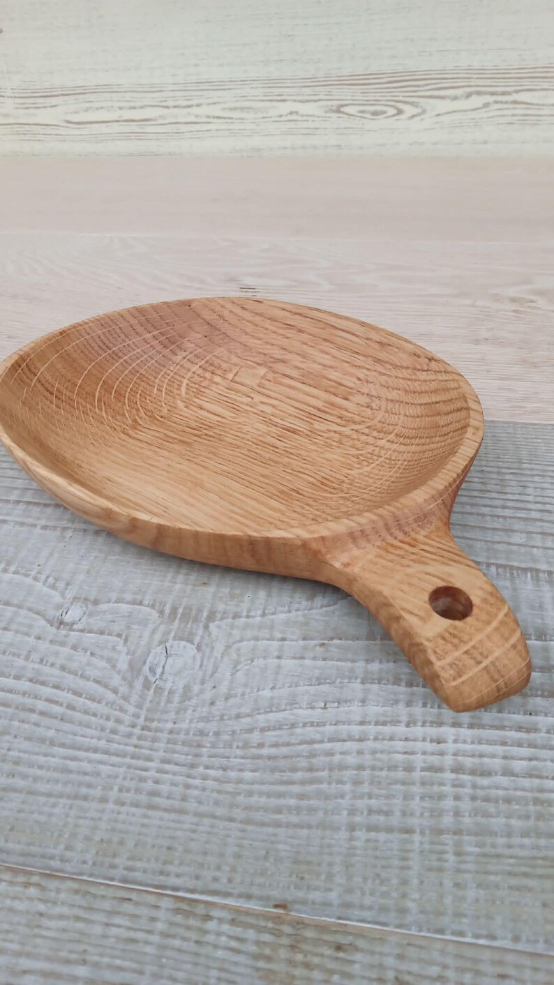 K&ouml;nnte beinhalten: Ein hellbraunes, ovales Serviertablett aus Holz mit Griff. Das Tablett hat eine flache Schale und einen Griff mit einem Loch zum Aufh&auml;ngen. Die Holzmaserung ist &uuml;ber das gesamte Tablett sichtbar, das etwa 25 cm lang ist.