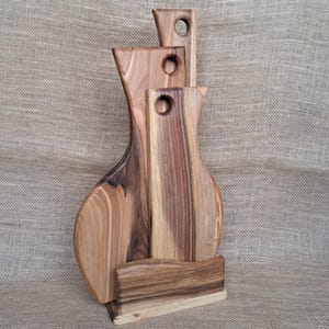 Op de afbeelding: Een set van drie houten snijplanken met natuurlijke houtnerven, elk voorzien van een rond gat aan de bovenkant om op te hangen. De planken staan verticaal opgesteld en worden vastgehouden door een bijpassende houten standaard met gleuf.