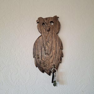 May include: A wooden owl-shaped key holder with a natural wood grain finish. The key holder has a carved design with two eye holes and a beak. Several hooks are attached to the bottom for hanging keys. A set of keys hangs from one of the hooks.