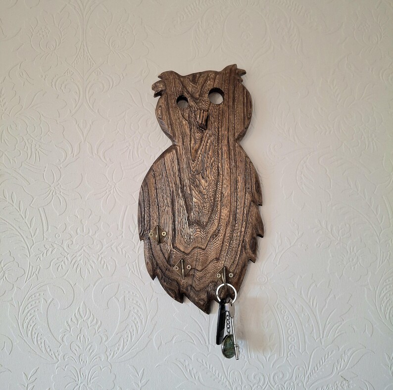 May include: A wooden owl-shaped key holder with a natural wood grain finish. The key holder has a carved design with two eye holes and a beak. Several hooks are attached to the bottom for hanging keys. A set of keys hangs from one of the hooks.
