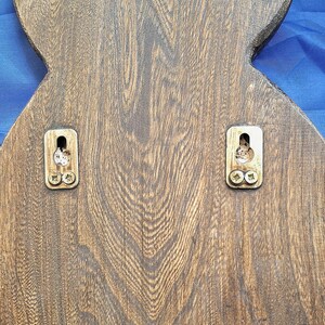 May include: Close-up of the back of a wooden decorative item, featuring a dark brown finish with visible wood grain. Two metal mounting brackets are attached with screws, ready for hanging. The item has a unique, possibly stylized shape.