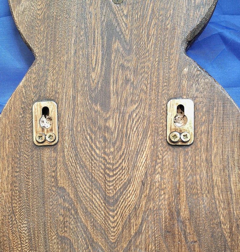 May include: Close-up of the back of a wooden decorative item, featuring a dark brown finish with visible wood grain. Two metal mounting brackets are attached with screws, ready for hanging. The item has a unique, possibly stylized shape.