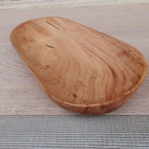 May include: A handcrafted wooden bowl with a natural, light brown finish. The bowl has a unique, elongated shape and visible wood grain patterns. The interior is smooth, and the rim is slightly raised. This decorative bowl is suitable for serving or display.