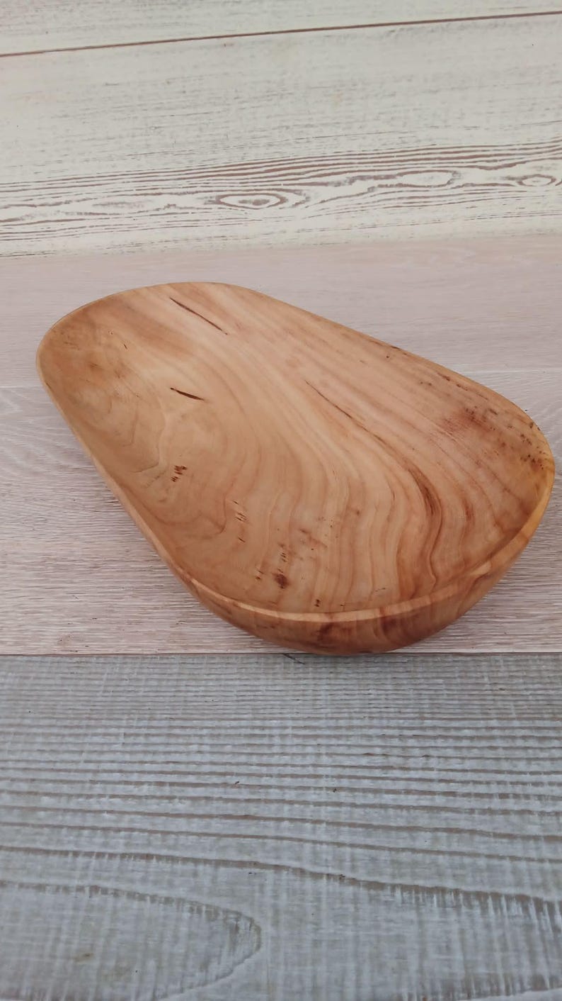 May include: A handcrafted wooden bowl with a natural, light brown finish. The bowl has a unique, elongated shape and visible wood grain patterns. The interior is smooth, and the rim is slightly raised. This decorative bowl is suitable for serving or display.
