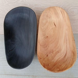 May include: Two wooden bowls, one black and one natural wood-toned, are displayed side-by-side. The bowls have an oblong shape with rounded edges. The black bowl has a smooth, dark finish, while the other bowl shows the natural wood grain.