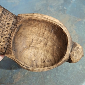 Pu&ograve; includere: Ciotola in legno intagliata a mano con finitura naturale. La ciotola presenta una forma arrotondata con un manico intagliato e dettagli decorativi. La venatura del legno &egrave; visibile, aggiungendo all'estetica rustica. Un pezzo unico per servire o esporre.