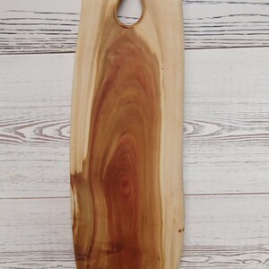May include: A tall, vertical wooden serving board with a natural light and dark brown wood grain pattern. The top section features two circular cutouts and a central notch, creating a shape that resembles the silhouette of an owl's head.