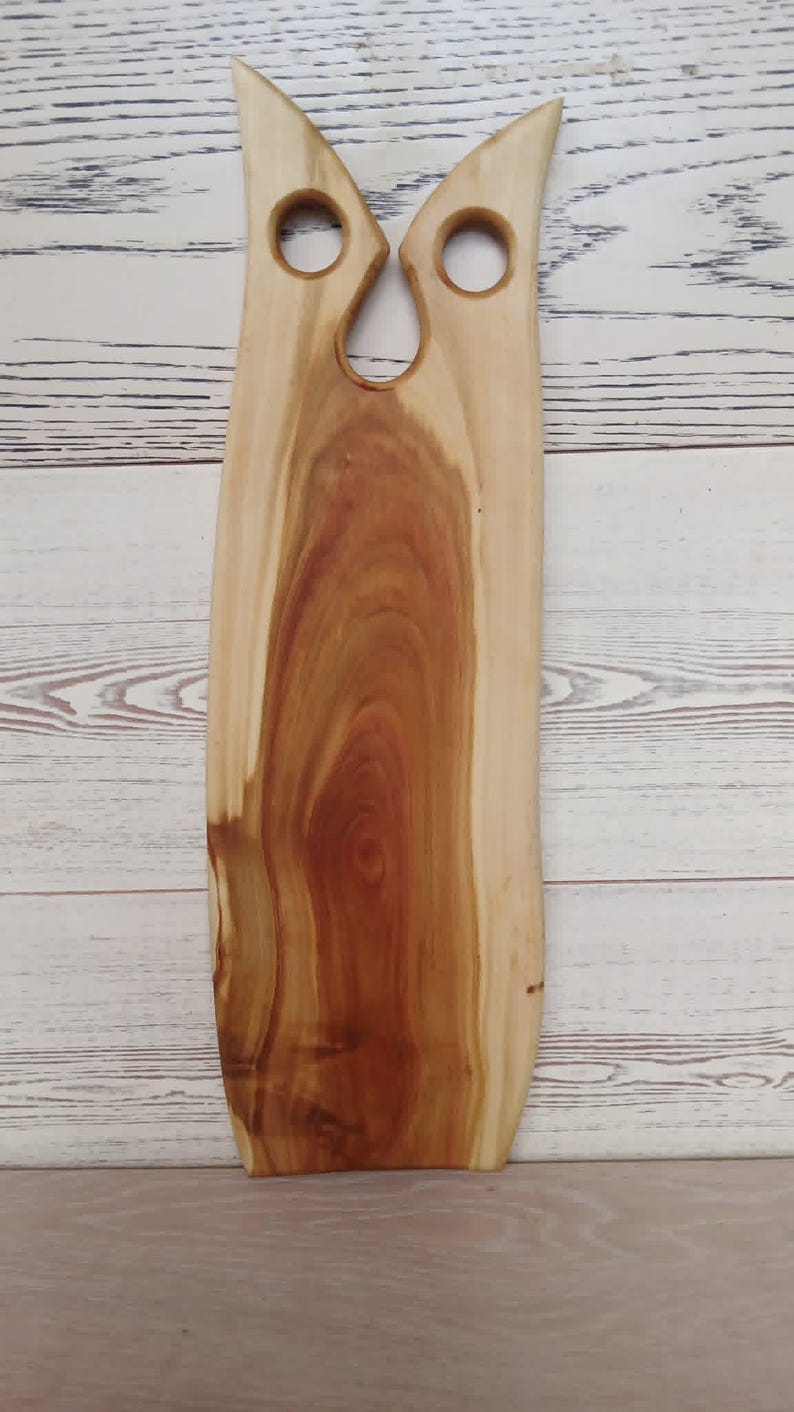 May include: A tall, vertical wooden serving board with a natural light and dark brown wood grain pattern. The top section features two circular cutouts and a central notch, creating a shape that resembles the silhouette of an owl's head.
