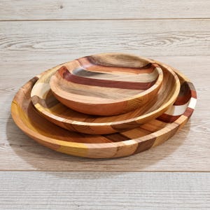 May include: A set of three nested circular wooden bowls stacked on top of each other. Each bowl features a multi-toned striped pattern created by joining different wood grains in shades of light tan, medium brown, and deep reddish-brown.