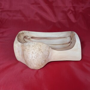 May include: A hand-carved wooden bowl with a natural finish. The bowl has a rectangular shape with rounded edges and a bulbous, textured detail on one side. The wood grain is visible throughout.