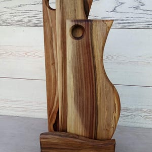 Op de afbeelding: Een set van drie verticale houten snijplanken met natuurlijke houtnerven en bruine tinten, elk voorzien van een rond gat aan de bovenkant om op te hangen. De planken staan rechtop in een bijpassende houten voet met een golvende voorzijde.