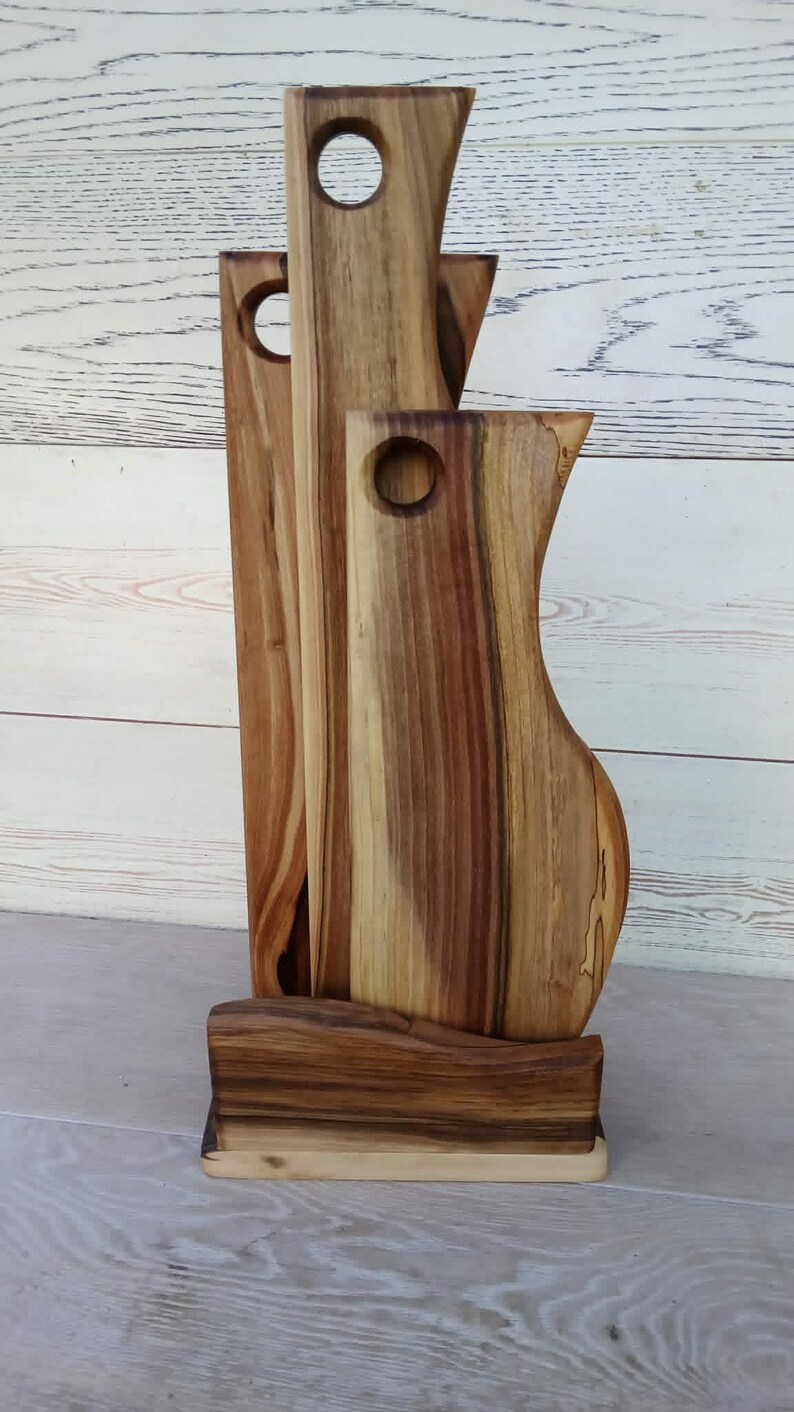 Op de afbeelding: Een set van drie verticale houten snijplanken met natuurlijke houtnerven en bruine tinten, elk voorzien van een rond gat aan de bovenkant om op te hangen. De planken staan rechtop in een bijpassende houten voet met een golvende voorzijde.