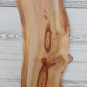 Puede incluir: Una tabla de servir de madera de color marr&oacute;n claro con un borde natural y un asa circular recortada en la parte superior. La tabla presenta patrones de grano de madera prominentes, incluyendo nudos en espiral y l&iacute;neas verticales.